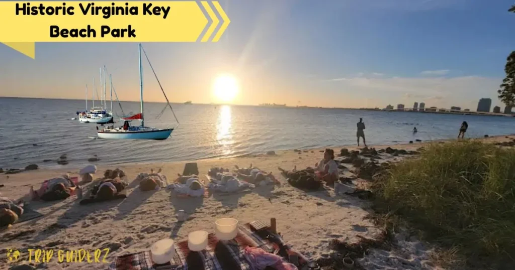 A serene sunset view at Historic Virginia Key Beach Park with sailboats on the water and people enjoying the calm bay