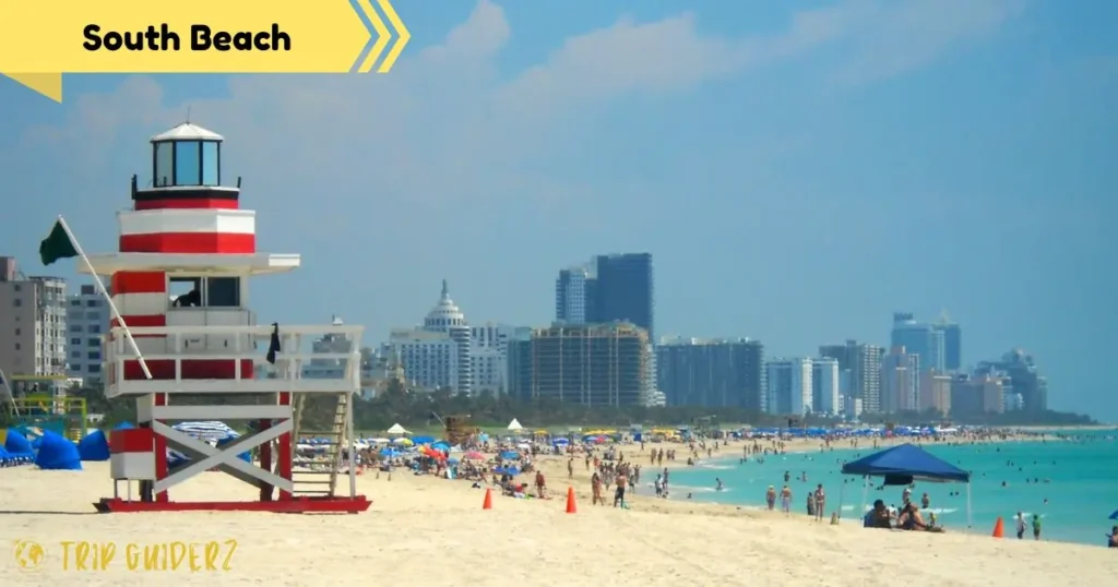 A wide view of a crowded South Beach with a classic red and white lifeguard stand in the foreground, showcasing a classic Miami Beach scene