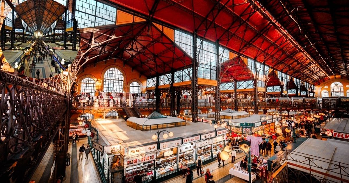 The interior of the Great Market Hall in Budapest, with vendors selling colorful produce and spices, and a wide view of the main floor and upper balconies.