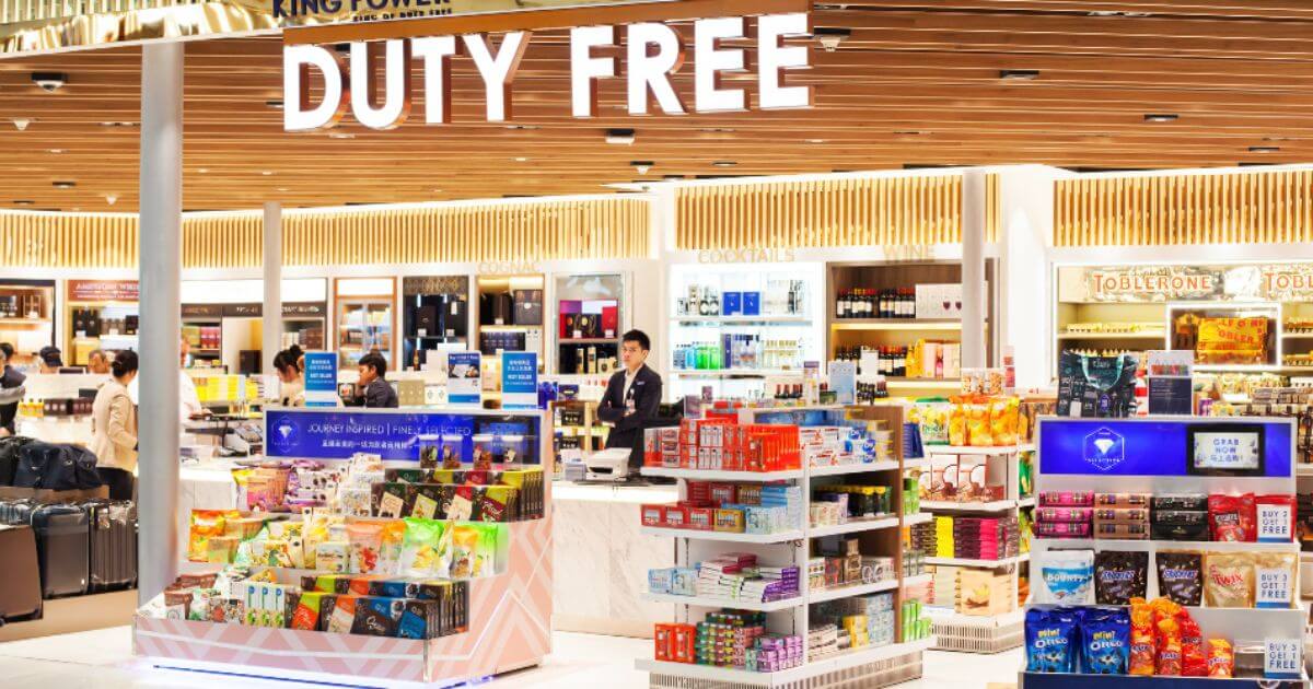 The interior of a modern, well-lit duty-free shop filled with shelves of various products like candy, liquor, and perfumes. The "DUTY FREE" sign is prominent above the entrance.