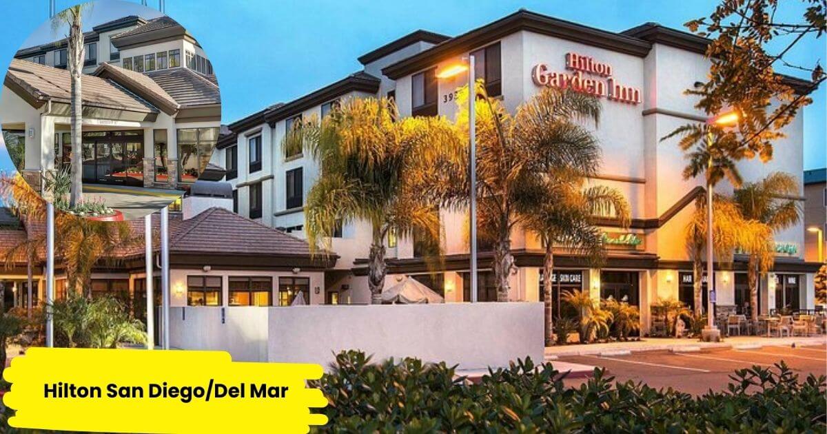 A nighttime photo of the Hilton Garden Inn San Diego/Del Mar, a hotel with a warm, inviting facade illuminated by exterior lighting. The building has a modern design with large windows and a sign for the hotel's on-site cafe.