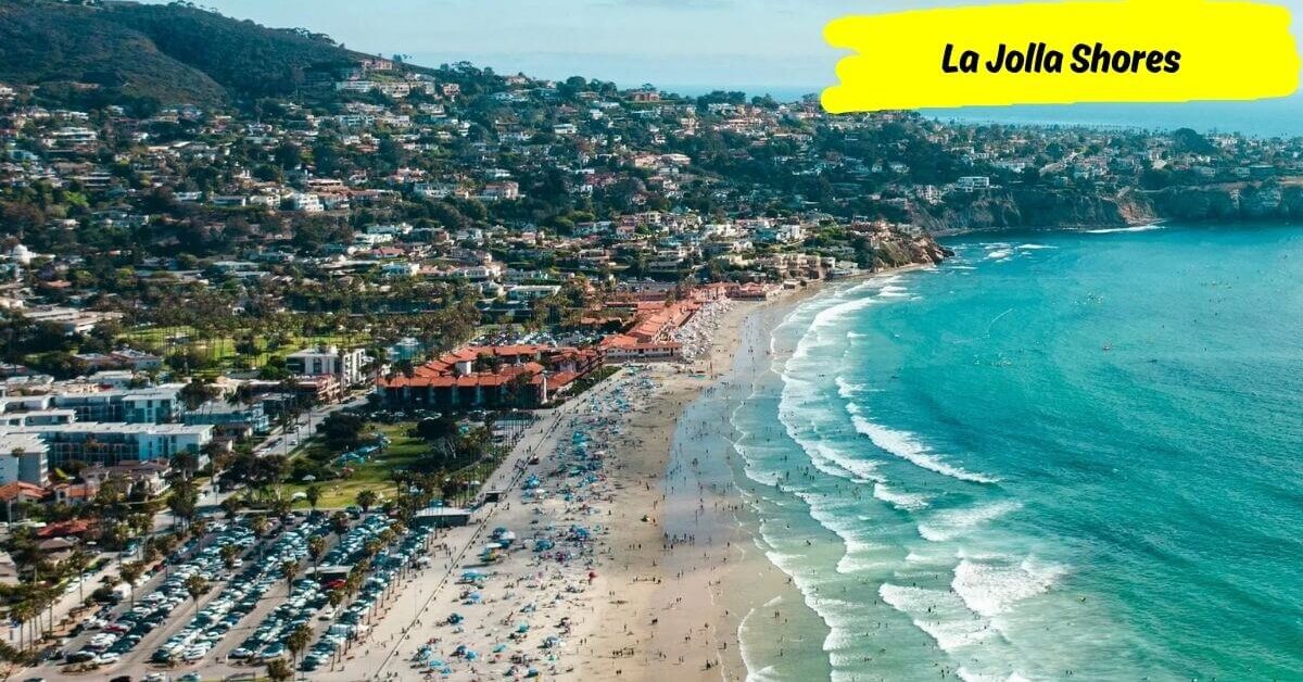 An aerial view of La Jolla Shores Beach showing a long, wide stretch of sand, surfers in the water, and homes on the hillside above.