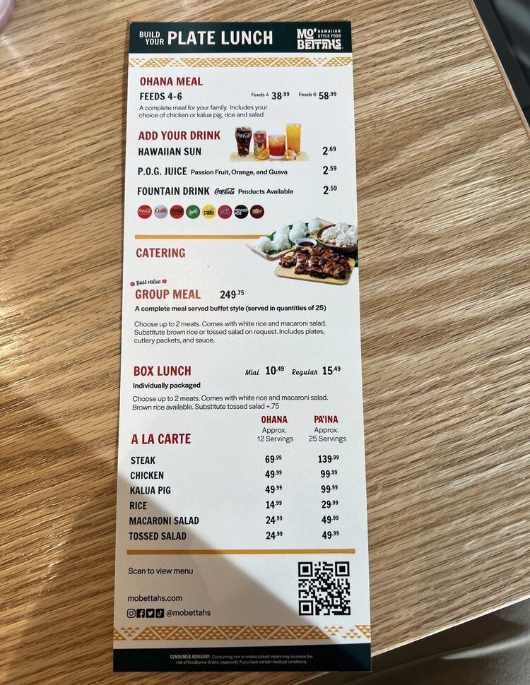 A close-up, vertical photo of a Mo' Bettahs "Build Your Plate Lunch" menu, detailing prices for Ohana Meals, Catering, Box Lunch options, and A La Carte items like Steak and Chicken. A QR code is visible at the bottom for scanning the menu.