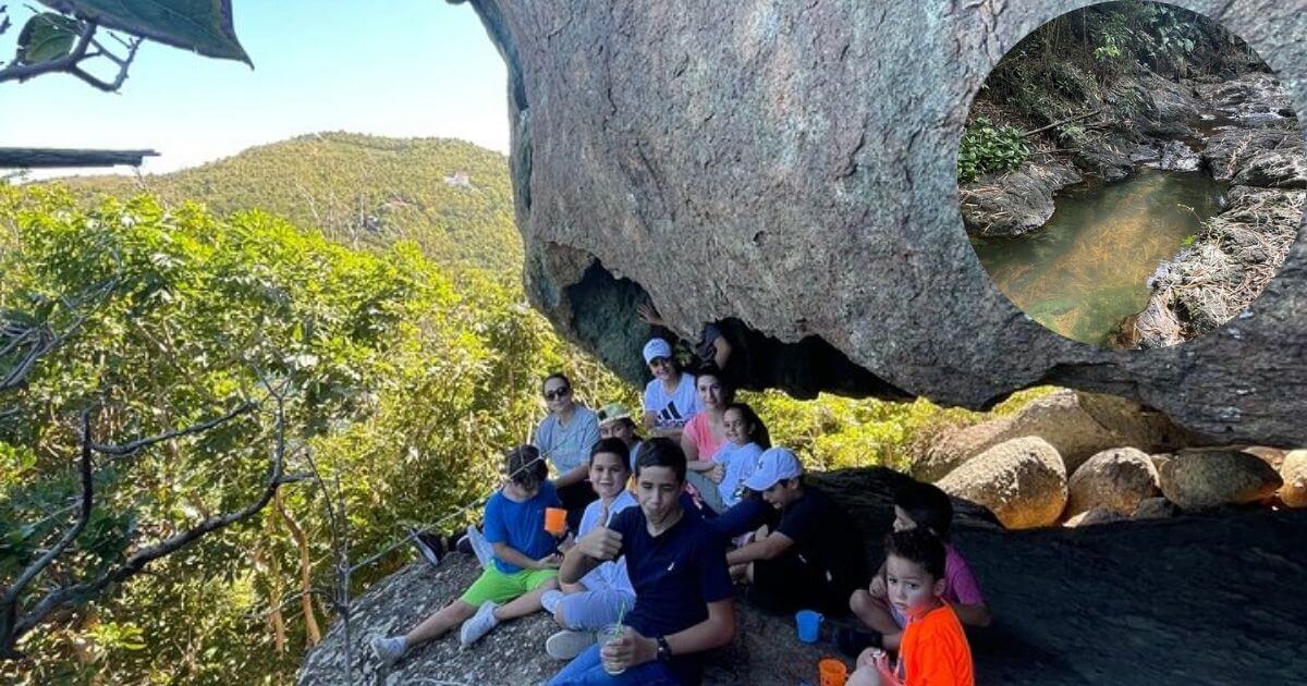 A group of hikers resting under a large boulder on a trail in Mount Healthy National Park, Tortola, with a scenic mountain view. An inset shows a small, rocky stream in the park.