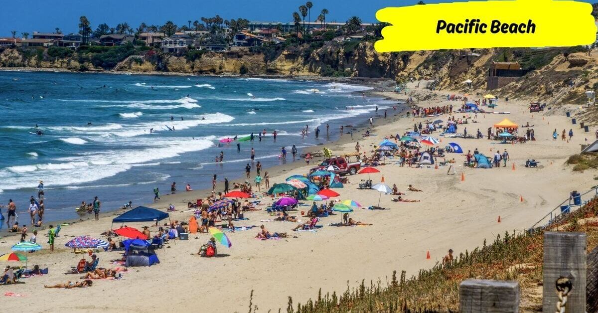 A sunny day at Pacific Beach with beachgoers, colorful umbrellas, and gentle waves rolling in, with cliffs in the background.