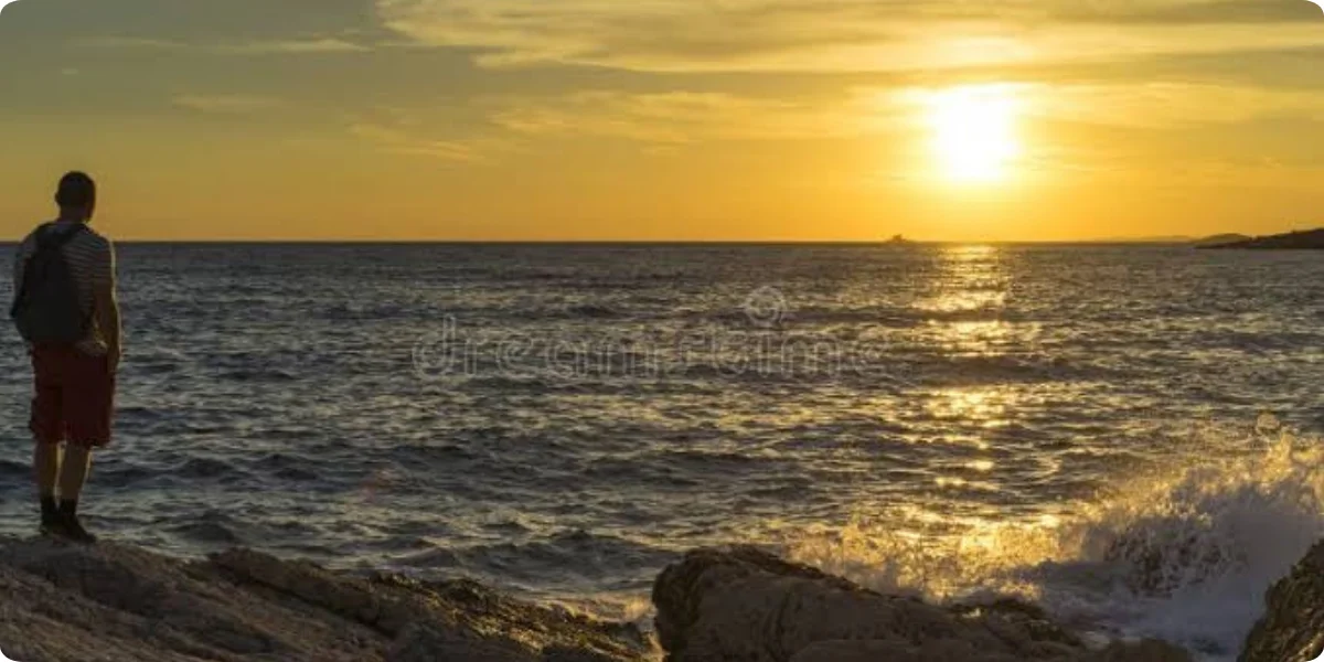 A man stands on rocky shore, gazing at a vibrant sunset reflecting on the water's surface.