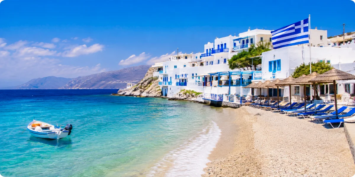 A serene Greek beach featuring blue chairs and a waving Greek flag against a clear sky.