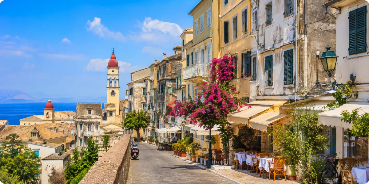 A narrow cobblestone street in Corfu's Old Town, lined with colorful buildings and vibrant flower pots.