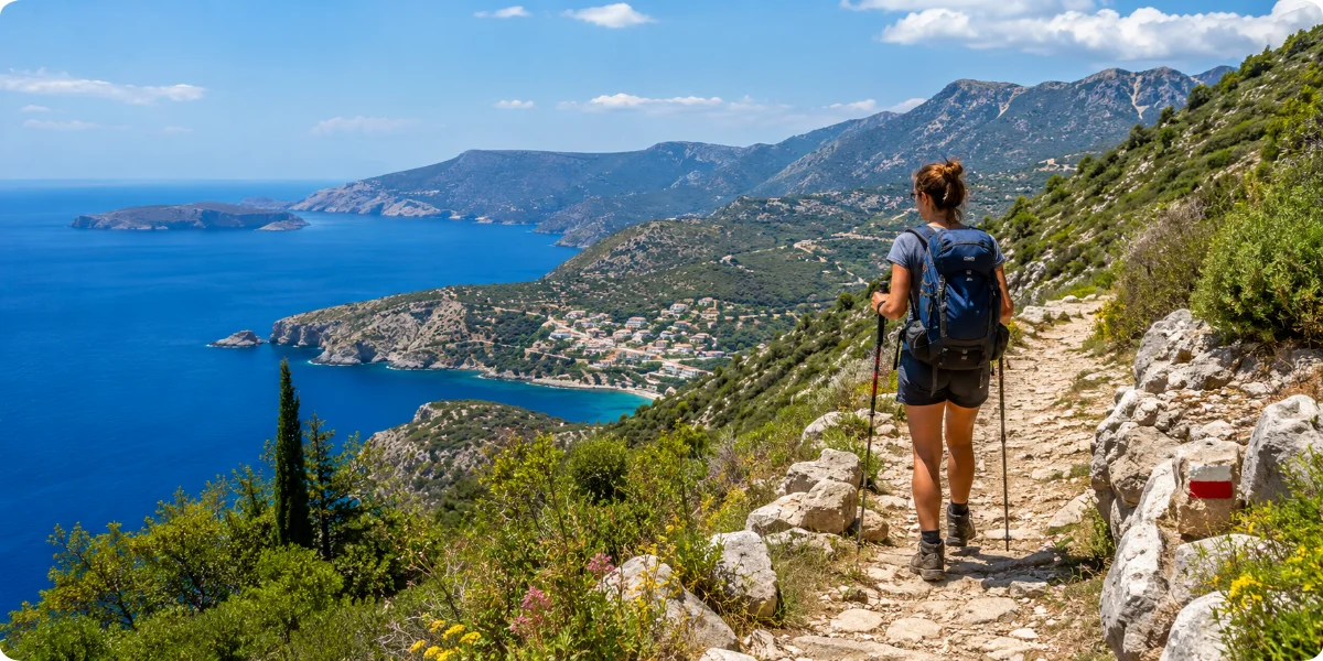 A woman traverses a scenic hiking trail in Greece, with the ocean visible in the background and vibrant nature surrounding her.