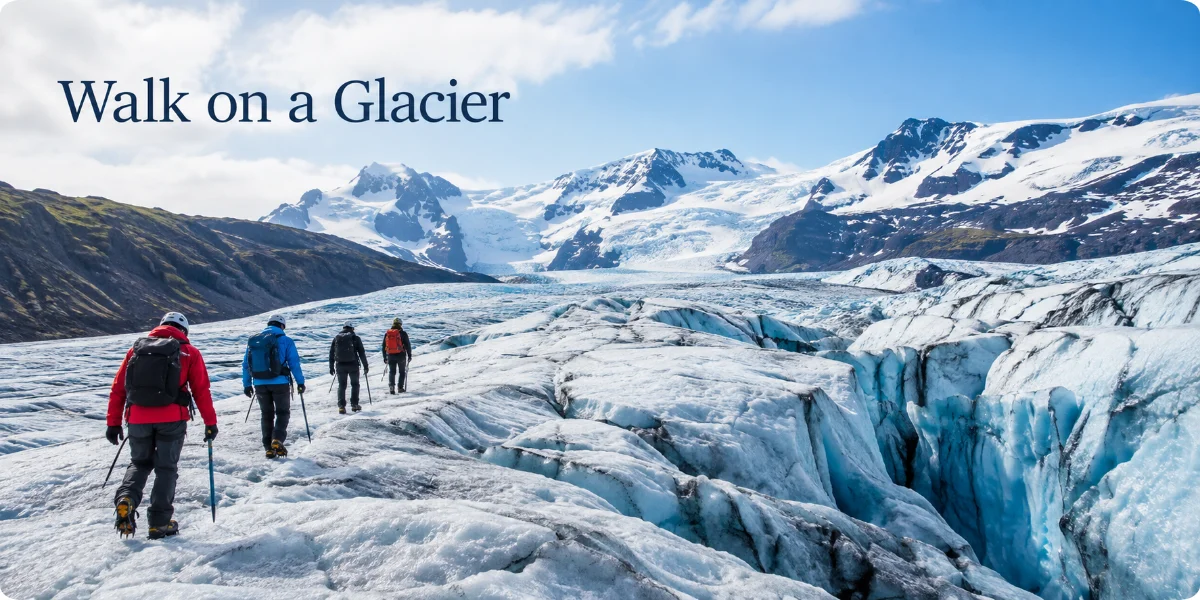 A person walking on a vast, icy glacier, surrounded by towering snow-covered mountains under a clear blue sky.